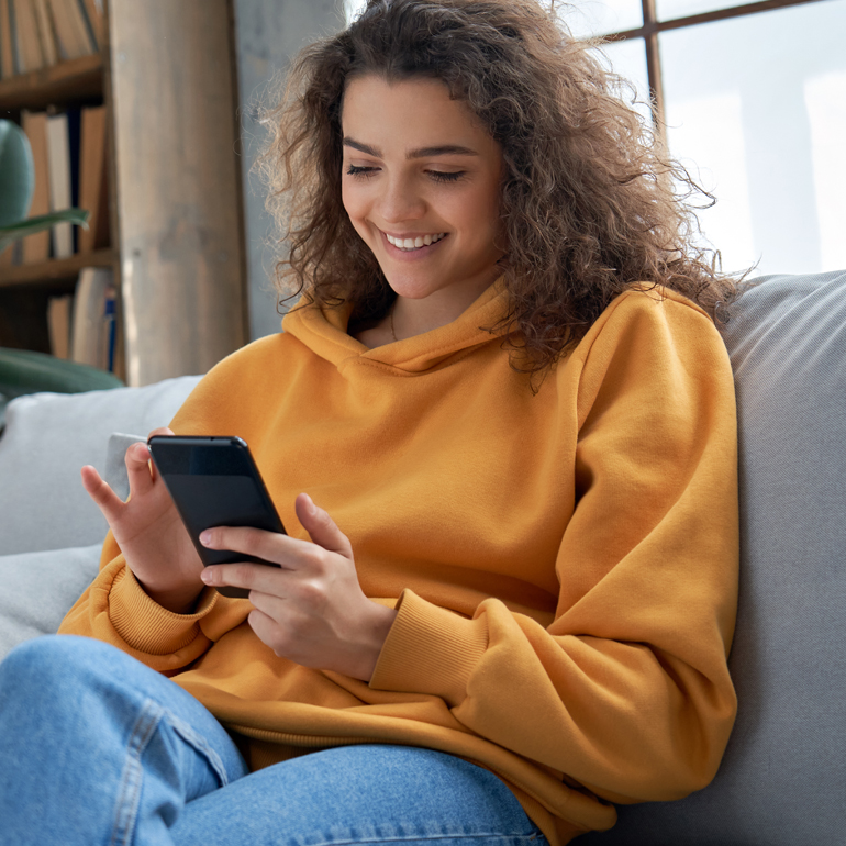 A person wearing an orange sweater and blue jeans sits on a couch using a smartphone.
